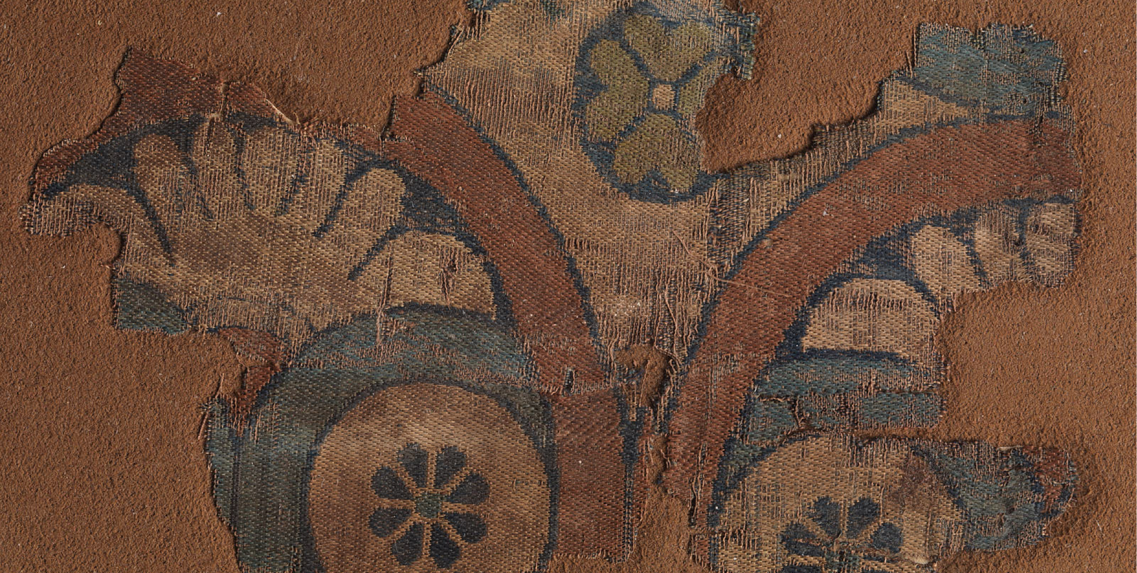 A fragment from a garment of silk samite with a twill ground. The fragment is patterned with partial floral roundels in gray, gold, and green on a brown ground.